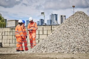 Three construction workers in orange safety gear review plans beside a large gravel pile at a city construction site with skyline in the background.