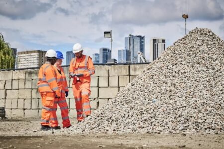 Three construction workers in orange safety gear review plans beside a large gravel pile at a city construction site with skyline in the background.