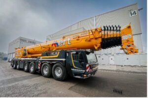 Large orange mobile crane mounted on a multi-axle truck parked beside a large industrial warehouse.