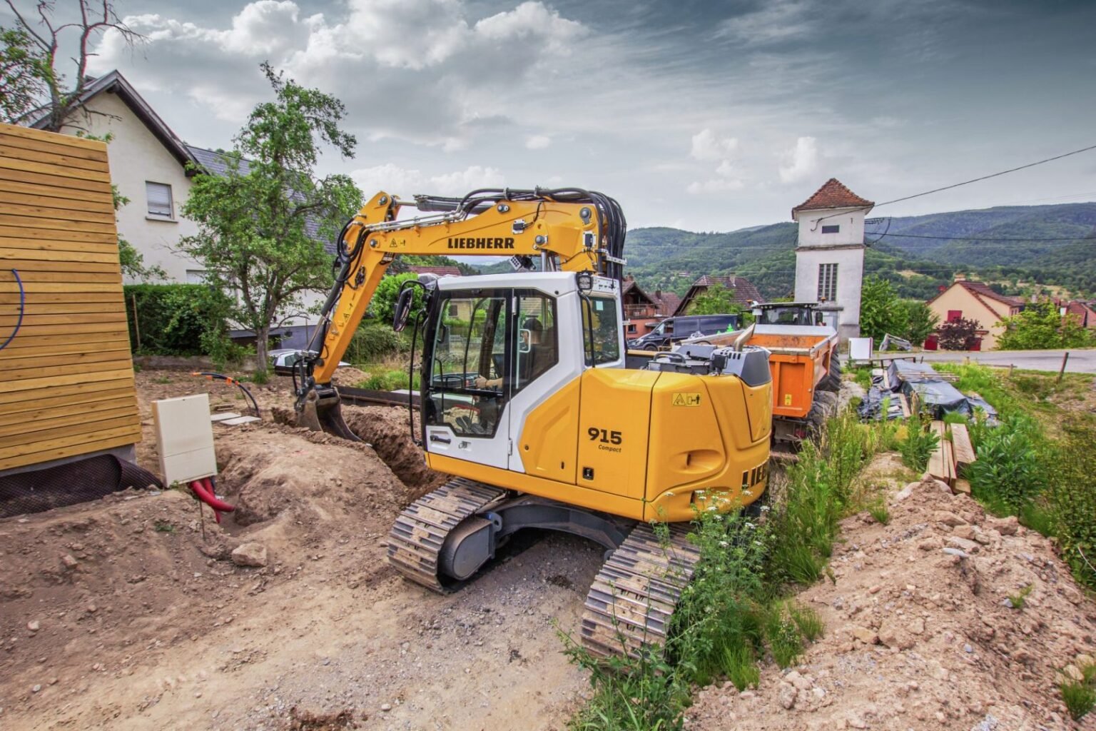 liebherr-r-915-compact-g8-300-dpi-scaledjpg - Machines Wiki Yellow Liebherr excavator working on a dirt trench at a residential construction site, with a village and hills in the background.
