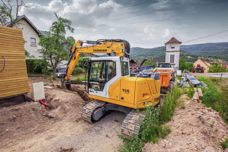 Yellow Liebherr excavator working on a dirt trench at a residential construction site, with a village and hills in the background.