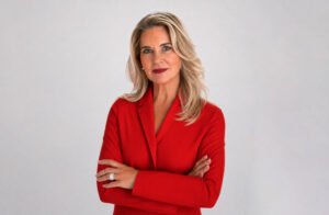 theresa-thomasjpg - Machines Wiki Professional woman with blonde hair in a red blazer, arms crossed, looking at the camera.