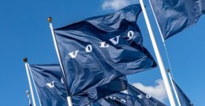 Blue flags with the 'VOLVO' wordmark fluttering on flagpoles against a clear blue sky.