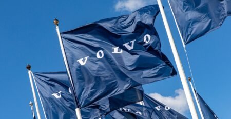 Blue flags with the 'VOLVO' wordmark fluttering on flagpoles against a clear blue sky.