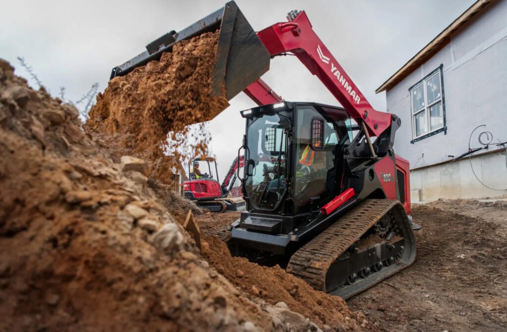 Large red excavator with a front bucket dumping dirt at a construction site beside a white building.