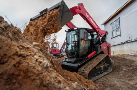 yanmar_jpg - Machines Wiki Large red excavator with a front bucket dumping dirt at a construction site beside a white building.