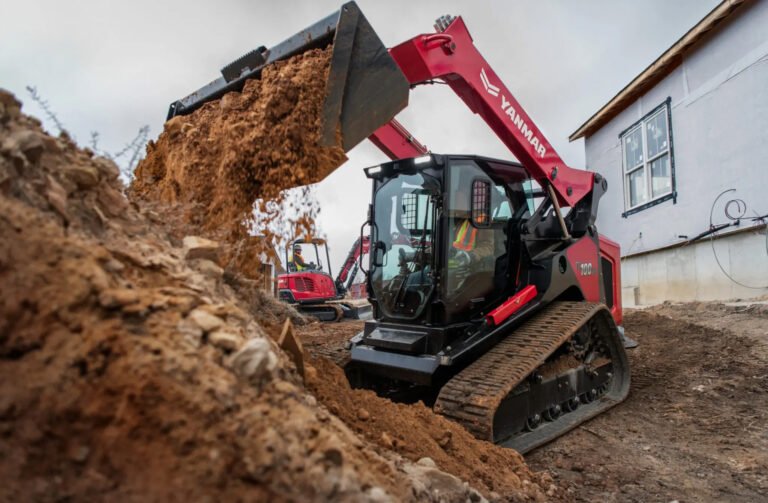 Large red excavator with a front bucket dumping dirt at a construction site beside a white building.