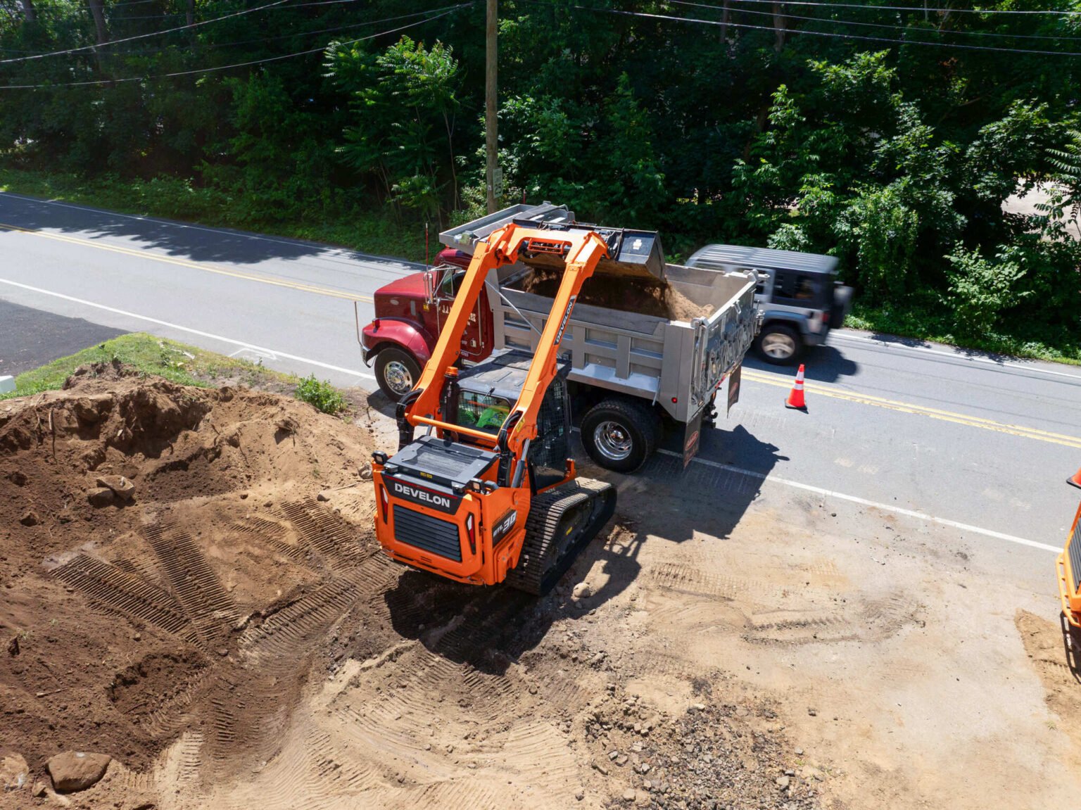 0d427401-f7a1-4c31-a8e8-ca89c7913710jpeg - Machines Wiki Construction site with an orange excavator unloading dirt into a dump truck beside a road. A trench is dug in the dirt foreground and traffic cones are visible along the street edge. Dense trees line the background.
