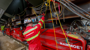 Worker in red safety suit and hard hat attaching yellow cables to a large red Mammoet machine at a construction site.