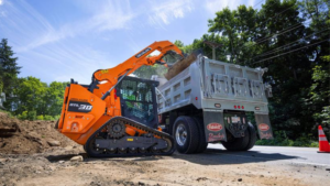 1785959040_64140_en_f547b_66320_develon-dtl30-compact-track-loaderpng - Machines Wiki Orange compact track loader transfers dirt into a gray dump truck on a roadside construction site, with trees and a traffic cone in the background.
