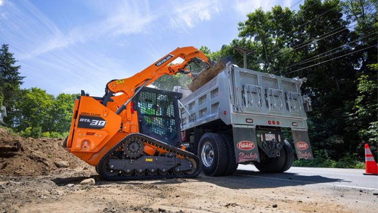 1785959040_64140_en_f547b_66320_develon-dtl30-compact-track-loaderpng - Machines Wiki Orange compact track loader transfers dirt into a gray dump truck on a roadside construction site, with trees and a traffic cone in the background.