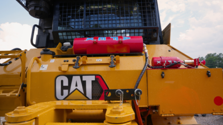 1786053021_64150_en_62c7f_66334_afex-3-gallon-fire-suppresion-systempng - Machines Wiki Close-up of a yellow CAT construction machine with a red fire extinguisher mounted on top and a protective grille ground-level view of the equipment