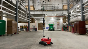 Red autonomous mobile robot on a warehouse floor at an industrial construction site, with scaffolding, large ducts, and workers in the background.
