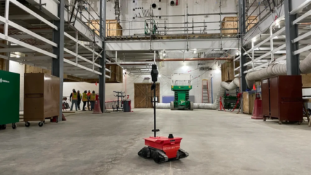 Red autonomous mobile robot on a warehouse floor at an industrial construction site, with scaffolding, large ducts, and workers in the background.