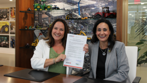 1786481616_64166_en_c46a9_66355_cga-aem-partnershippng - Machines Wiki Two women in a meeting room smile while presenting an official certificate; shelves of toy tractors and equipment in the background.