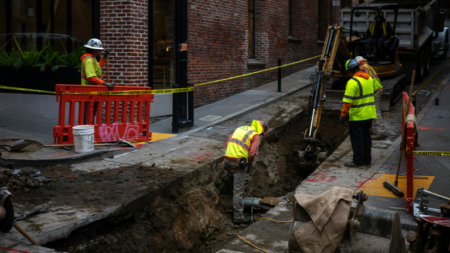Construction workers in high-visibility vests and hard hats excavate a trench along a city sidewalk, surrounded by safety barriers, yellow tape, and heavy equipment.