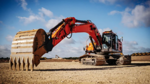 Red hydraulic excavator with a large digging bucket on a sandy construction site under a blue sky