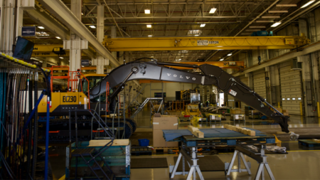 1787177914_64191_en_ed875_66382_volvo-ce-excavator-shippensburg-pennsylvaniapng - Machines Wiki Inside a manufacturing workshop, a Volvo hydraulic excavator arm is extended over a worktable with pallets nearby.