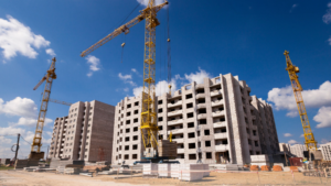 Construction site with multiple unfinished gray concrete buildings and three yellow cranes under a clear blue sky, indicating ongoing multi-story construction.