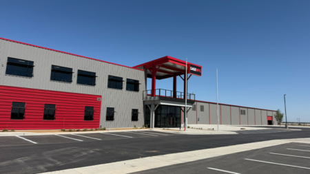 Exterior of a gray industrial building with red accents and a red canopy over the main entrance; empty parking lot under a blue sky.