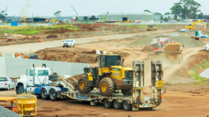 1787689258_64224_en_dd228_66419_unsplash-heavy-equipmentpng - Machines Wiki Construction site with a yellow front-end loader on a flatbed trailer, being towed by a white truck, amid dirt and other heavy equipment.