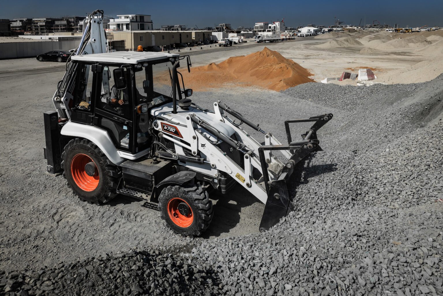 White compact front-end loader with orange wheels on a gravel construction site, with piles of sand and distant buildings in the background.