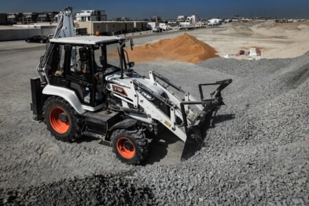 20251114_Bobcat_CustomerStories_Dubai_Day5_2503_redjpg - Machines Wiki White compact front-end loader with orange wheels on a gravel construction site, with piles of sand and distant buildings in the background.