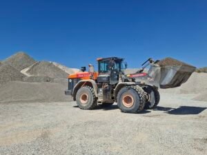 Orange heavy-duty dump truck at a gravel quarry, with large piles of crushed rock and a clear blue sky in the background.