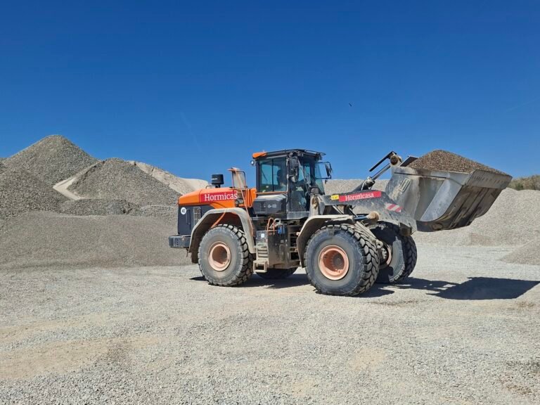 Orange heavy-duty dump truck at a gravel quarry, with large piles of crushed rock and a clear blue sky in the background.