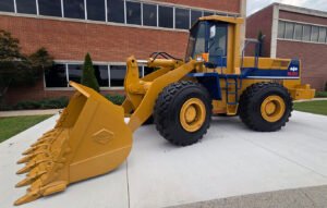 Yellow WA450 wheel loader with a large toothed bucket, parked on a concrete pad in front of a brick building.