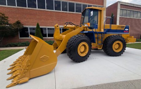 20260710_102817jpg - Machines Wiki Yellow WA450 wheel loader with a large toothed bucket, parked on a concrete pad in front of a brick building.