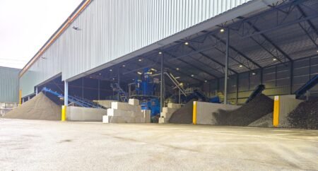 Industrial warehouse with blue machinery and conveyors handling gravel piles inside a large open-sided shed