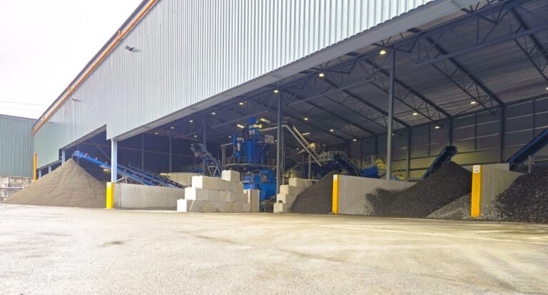 Industrial warehouse with blue machinery and conveyors handling gravel piles inside a large open-sided shed