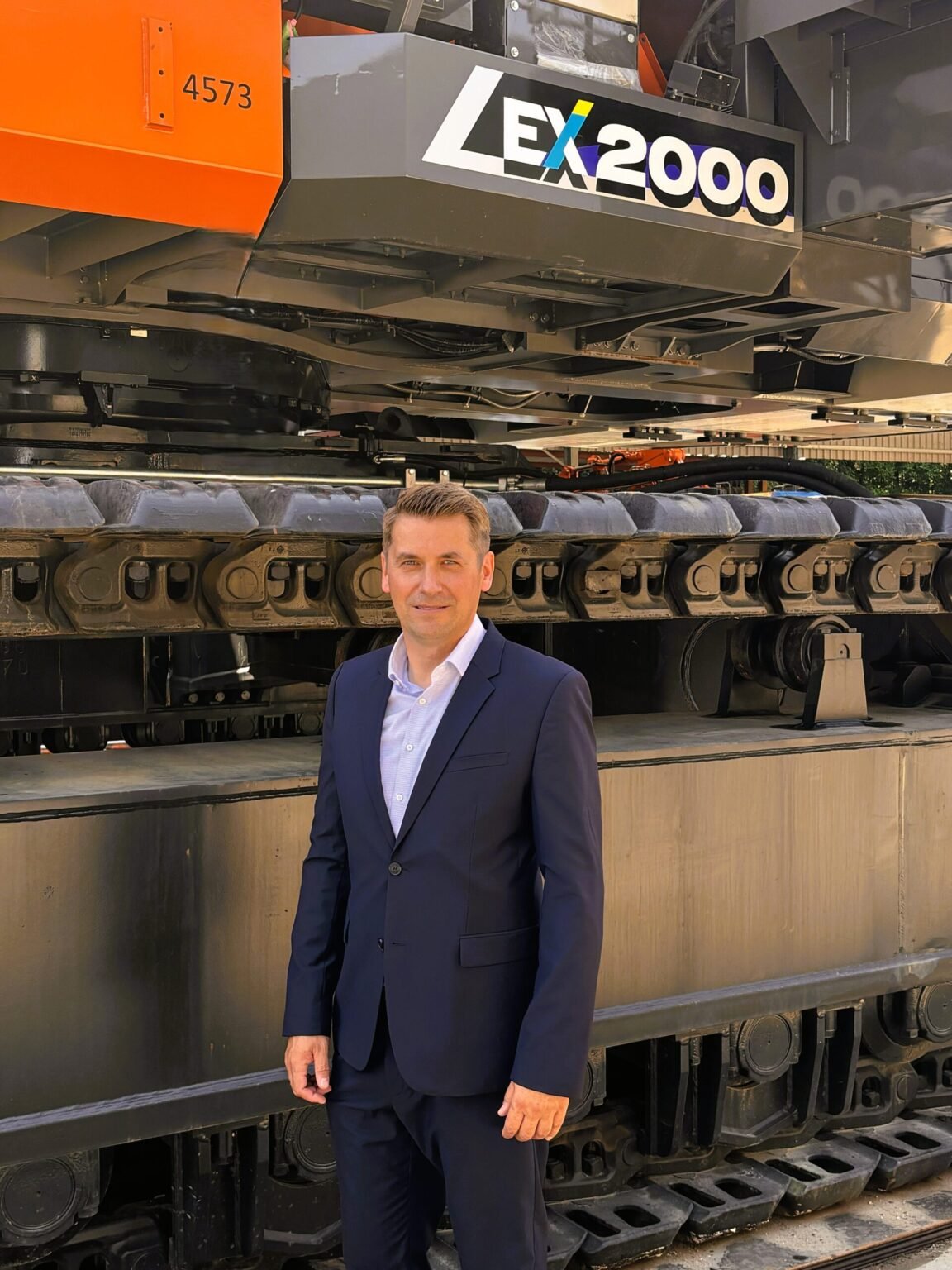 HM_June-2026-1-1-scaledjpg - Machines Wiki Man in a dark blue suit standing beside a large industrial machine with orange panel and exposed tracks labeled EX2000/4573 in the background.