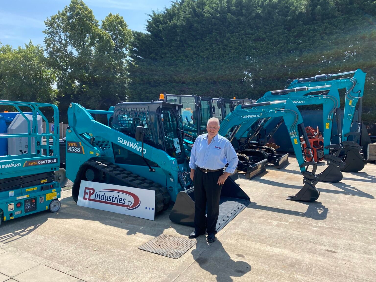 IMG_082-scaledjpg - Machines Wiki Smiling man in a light blue shirt stands among turquoise Sunward excavators at a construction equipment yard, with a banner reading EP Industries Ltd nearby.
