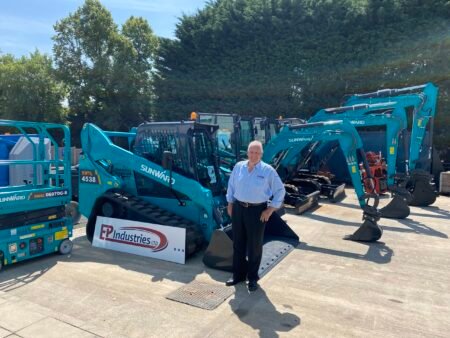 IMG_082-scaledjpg - Machines Wiki Smiling man in a light blue shirt stands among turquoise Sunward excavators at a construction equipment yard, with a banner reading EP Industries Ltd nearby.