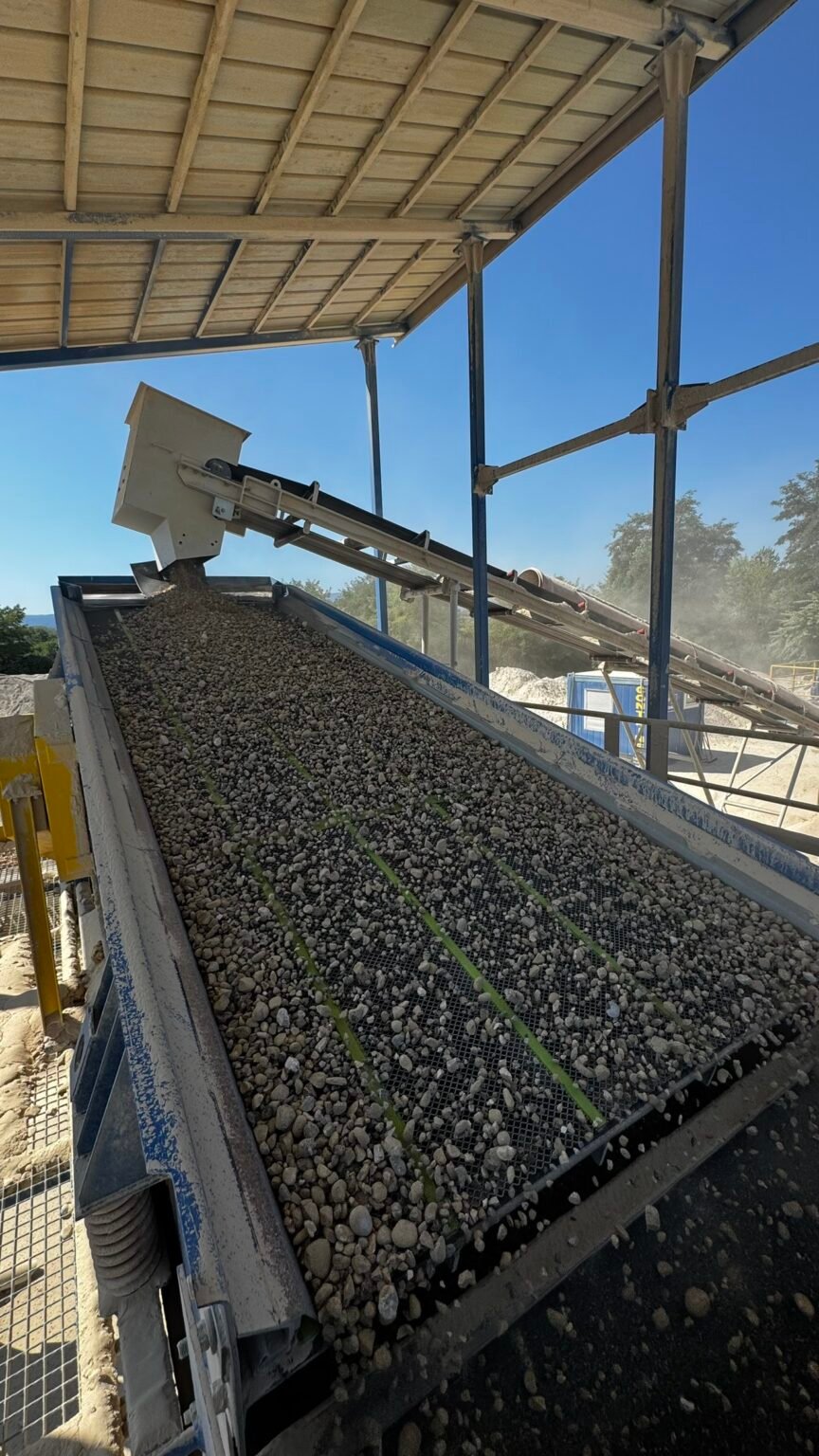 MAJOR_steinexpo_1jpeg - Machines Wiki Conveyor belt dumping a load of gravel under a metal-framed shelter at a construction site.