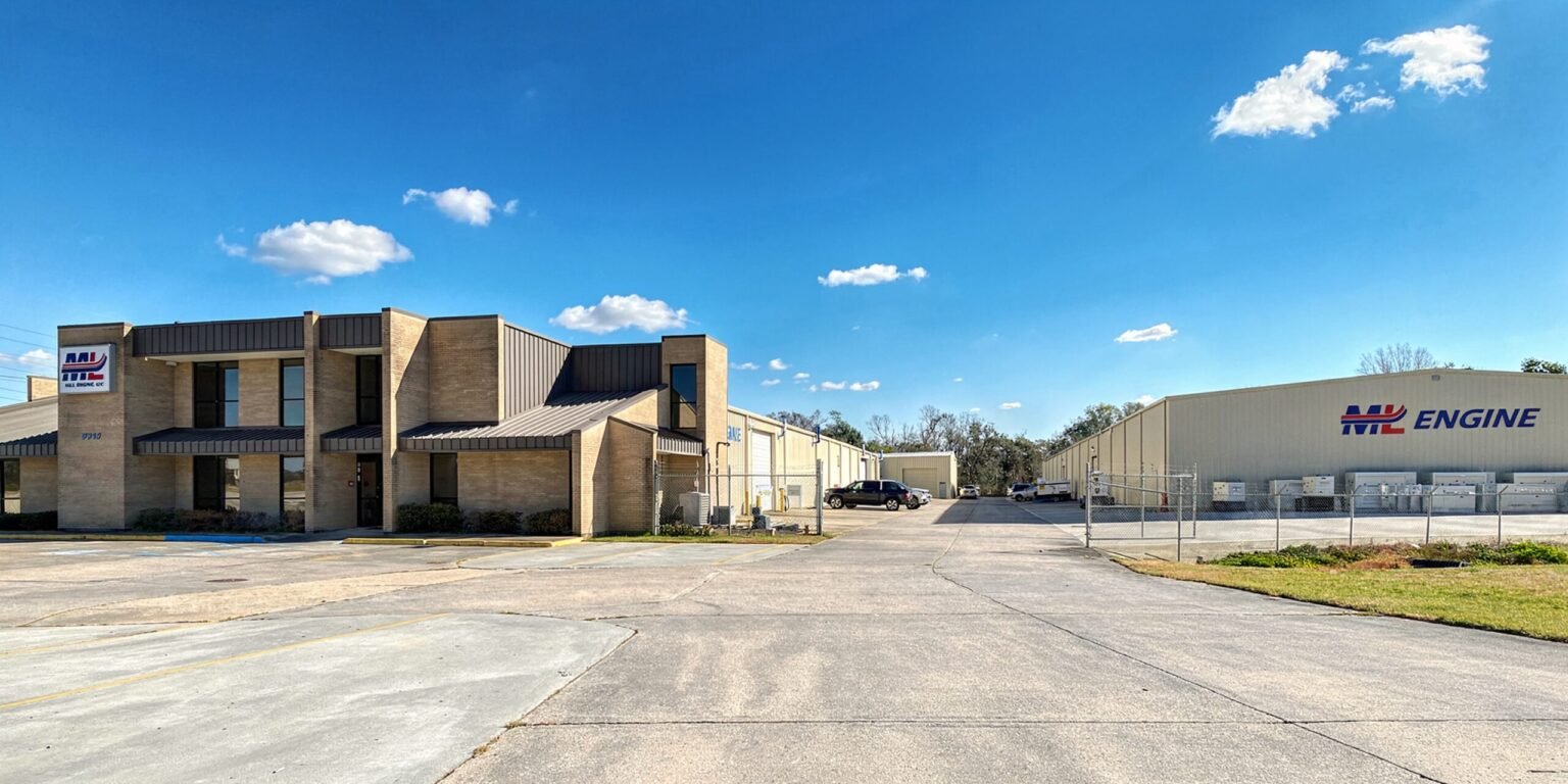 ML-Engine-Facility-2026-A_edited_new-scaledjpg - Machines Wiki Exterior view of a brick office building beside a large cream-colored industrial warehouse under a clear blue sky with a fence in front and parked cars nearby.