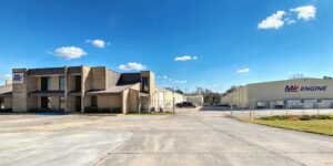 FPT Announces the Expansion of M&L Engine Partnership into the Carolinas, in the U.S. Exterior view of a brick office building beside a large cream-colored industrial warehouse under a clear blue sky with a fence in front and parked cars nearby.