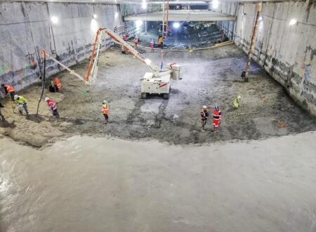 Underground construction site with a muddy floor; workers in safety vests and helmets operate a concrete pump truck.