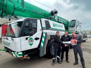 Meijmat-modernizes-crane-fleet-with-new-Grove-GMK5120L-all-terrain-crane-01jpg - Machines Wiki Three workers in dark uniforms pose in front of a large white-and-green Meumat crane truck with a bright green boom in a parking lot.