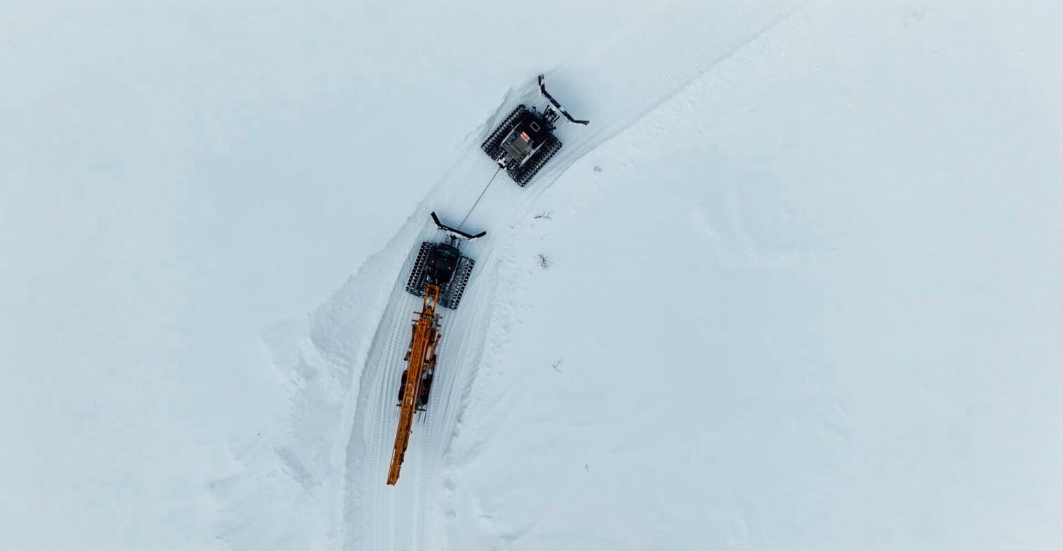 Potain-self-erecting-crane-reaches-new-heights-on-Norway-mountaintop-project-02jpg - Machines Wiki Aerial view of an orange crane with a long boom on a snow-covered field, tethered to a second tracked vehicle.