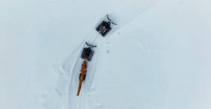 Potain-self-erecting-crane-reaches-new-heights-on-Norway-mountaintop-project-02jpg - Machines Wiki Aerial view of an orange crane with a long boom on a snow-covered field, tethered to a second tracked vehicle.