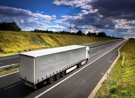Senza-titolo-22-giugno-2026-alle-ore-153503-2-scaledjpeg - Machines Wiki White semi-trailer truck driving on a highway beside a grassy embankment under a blue sky with clouds and power lines nearby.