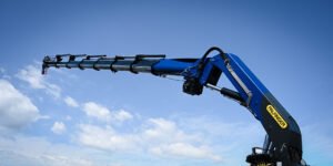 The-new-PFM-1200-impresses-with-its-outreach-performance-and-durability-jpg - Machines Wiki Blue Palfinger crane arm extended horizontally against a clear blue sky with exposed hydraulics and hook at the end.