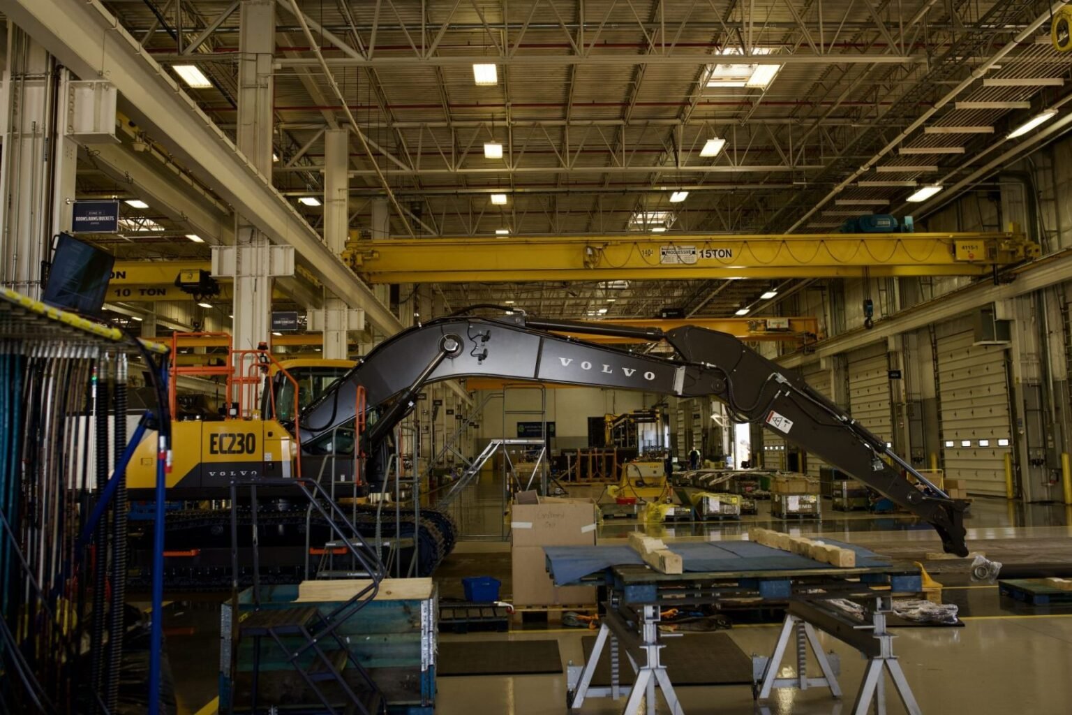 Volvo-CE-factory-image-scaledjpeg - Machines Wiki Industrial warehouse scene with a large Volvo EC230 excavator under a yellow overhead crane, surrounded by workbenches, pallets and tools.