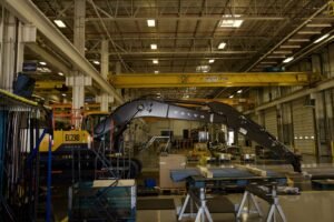 Industrial warehouse scene with a large Volvo EC230 excavator under a yellow overhead crane, surrounded by workbenches, pallets and tools.