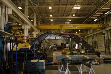 Volvo-CE-factory-image-scaledjpeg - Machines Wiki Industrial warehouse scene with a large Volvo EC230 excavator under a yellow overhead crane, surrounded by workbenches, pallets and tools.