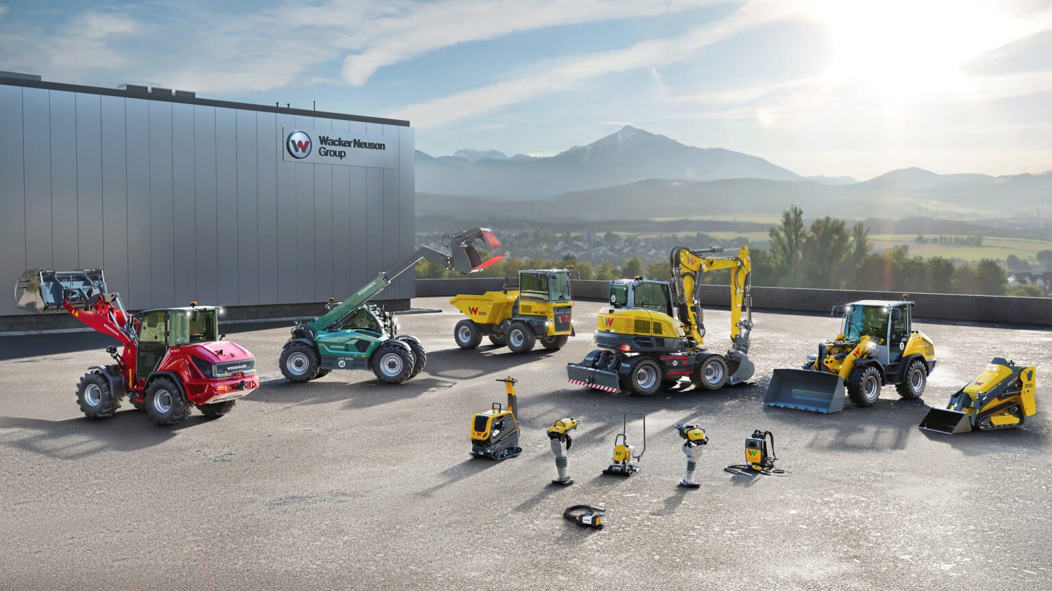 Industrial yard with yellow, red, and green construction machines parked beside a building bearing the Wacker Neuson Group sign; mountains in the background.