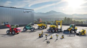 Wacker-Neuson-Group_H1jpg - Machines Wiki Industrial yard with yellow, red, and green construction machines parked beside a building bearing the Wacker Neuson Group sign; mountains in the background.